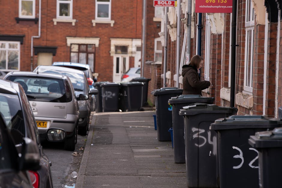 A sanitation worker wearing a high-visibility yellow and red uniform is engaged in rubbish collection next to a large red waste collection vehicle parked on the side of a road. The rear of the truck f