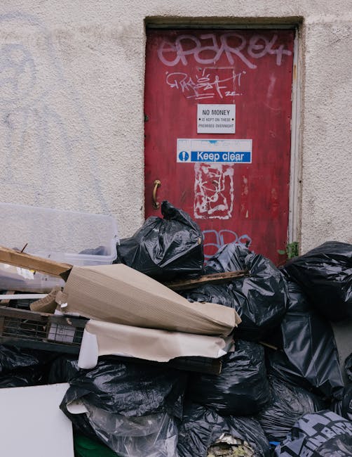 An exterior scene showing a rough, textured beige wall with a small opening above a red-painted steel door, which is covered in graffiti tags and stickers, including a prominent white and blue sign th