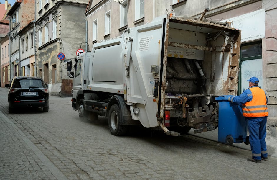 An urban scene showing an overflowing public rubbish bin on a paved sidewalk, surrounded by scattered household waste including cardboard boxes, plastic bags, paper, and packaging materials. The large