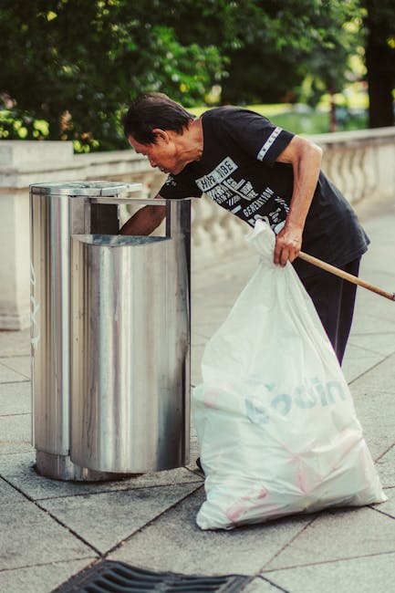 A black wheelie bin labeled 'ST. JOHN'S' sits on the pavement beside a residential street at night, with its lid open and filled with various waste items, including cardboard and plastic. The bin's su